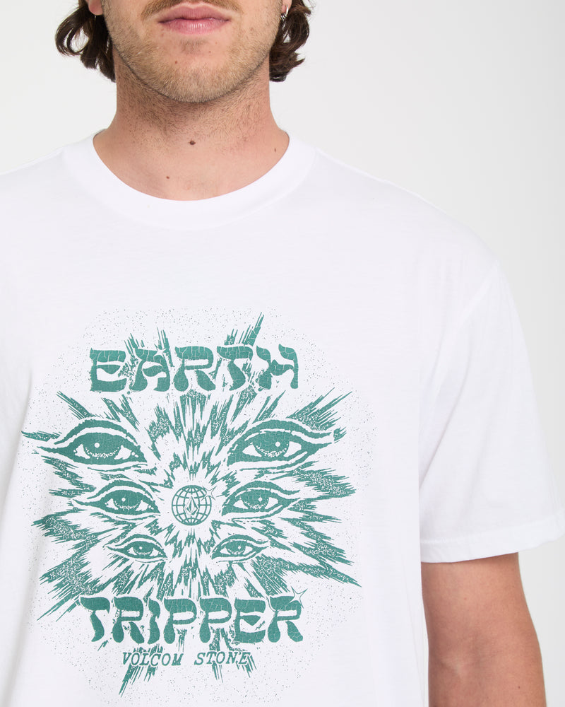 A man wears the Volcom Men's Remote Viewer Short Sleeve Tee in white, featuring a teal graphic with illustrated eyes, an Earth globe, and the text "EARTH TRIPPER" and "VOLCOM STONE." Only his lower face and upper torso are visible.