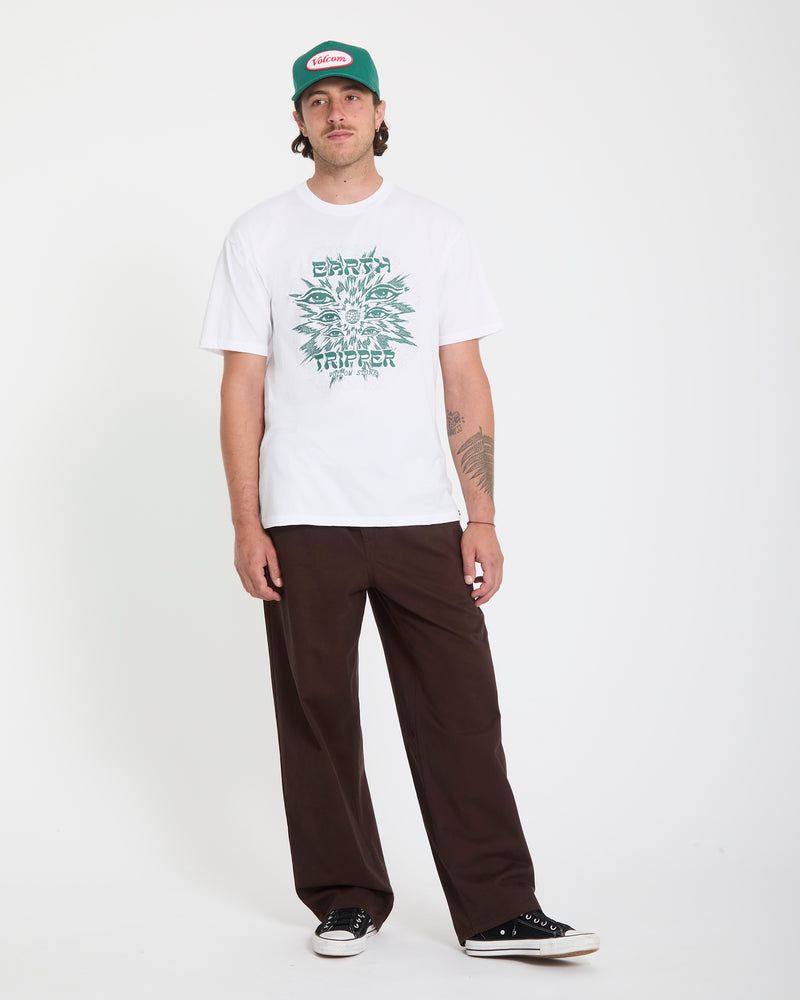 A man in a Volcom Men's Remote Viewer Short Sleeve Tee, green and white baseball cap, dark brown pants, and black sneakers stands against a plain white background.