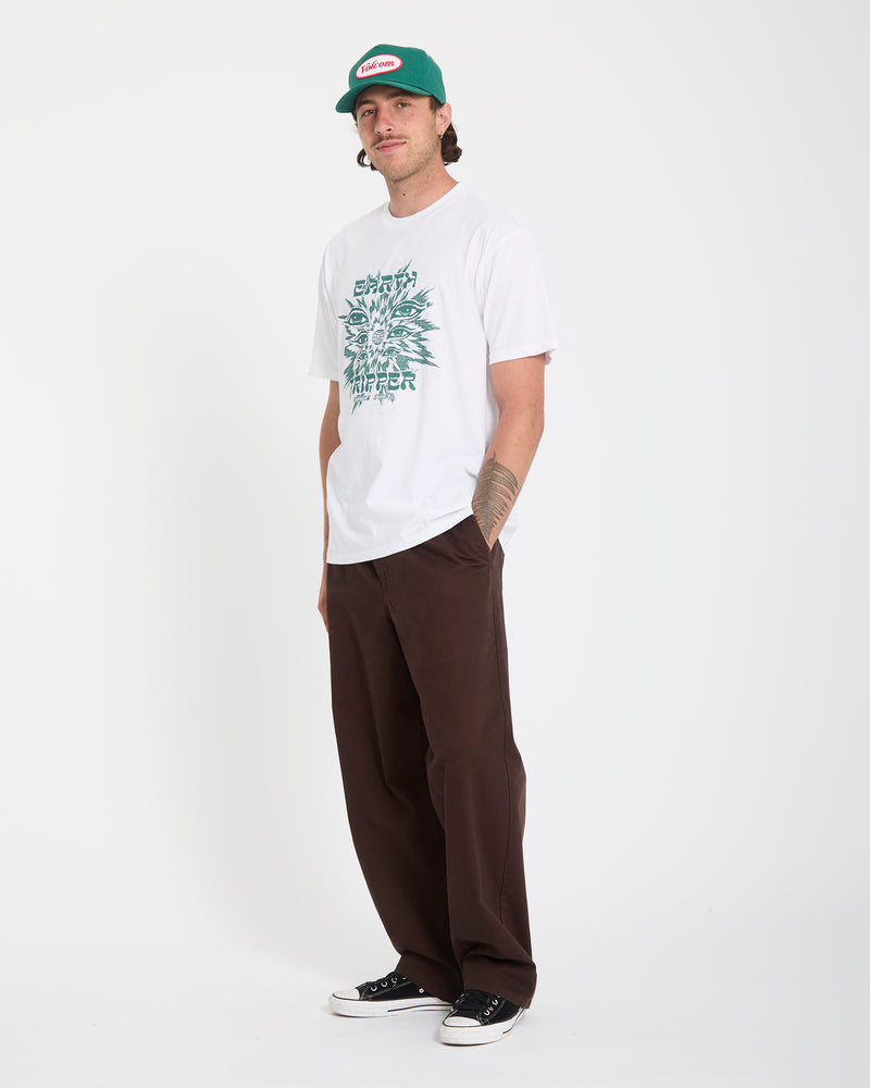 A man wears the Volcom Men's Remote Viewer Short Sleeve Tee in white, brown pants, black sneakers, and a green cap, standing with hands in pockets against a plain white background.