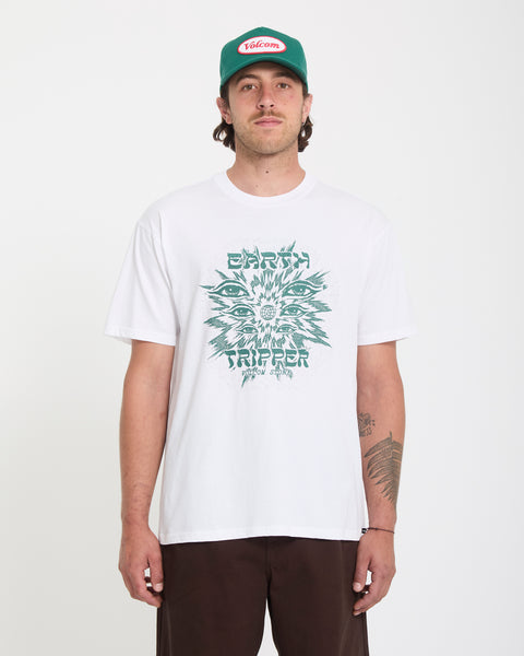A man faces forward wearing the Volcom Men's Remote Viewer Short Sleeve Tee in white with a green graphic and "Earth Tripper" text. He also sports a green and red cap, dark pants, and has a tattoo on his left forearm.