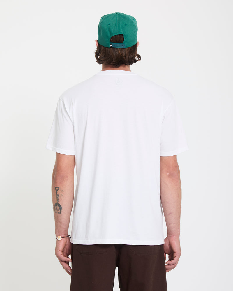 A man faces away in a Volcom Men's Remote Viewer Short Sleeve Tee (white), brown pants, and a green baseball cap. A tattoo and bracelet are visible on his left arm against a plain white background.