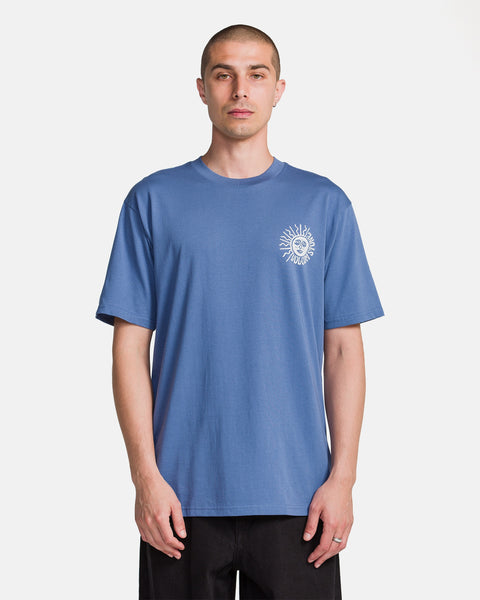 A man with short hair wears the Volcom Men's Mazatlan Short Sleeve Tee, a blue T-shirt with a small white sun graphic on the left chest, standing against a plain white background.