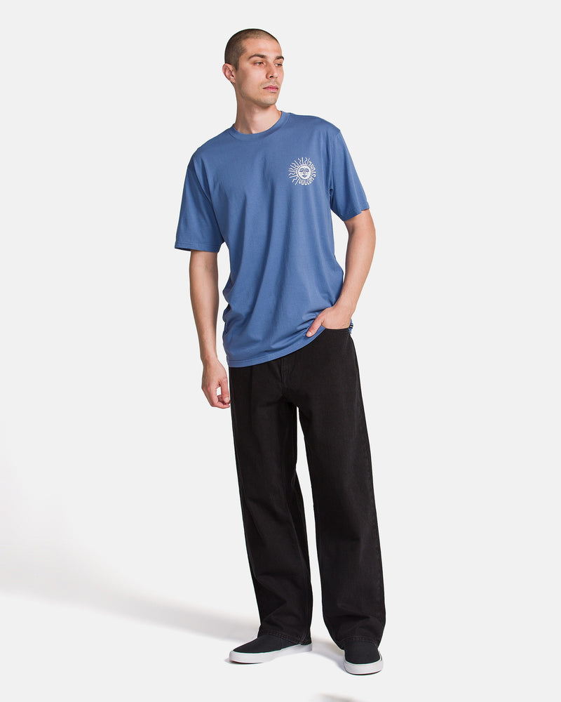 A person stands against a white background, wearing the Volcom Men's Mazatlan Short Sleeve Tee in blue with a small white chest graphic, paired with black pants and dark shoes. One hand is in their pocket as they look to the side.