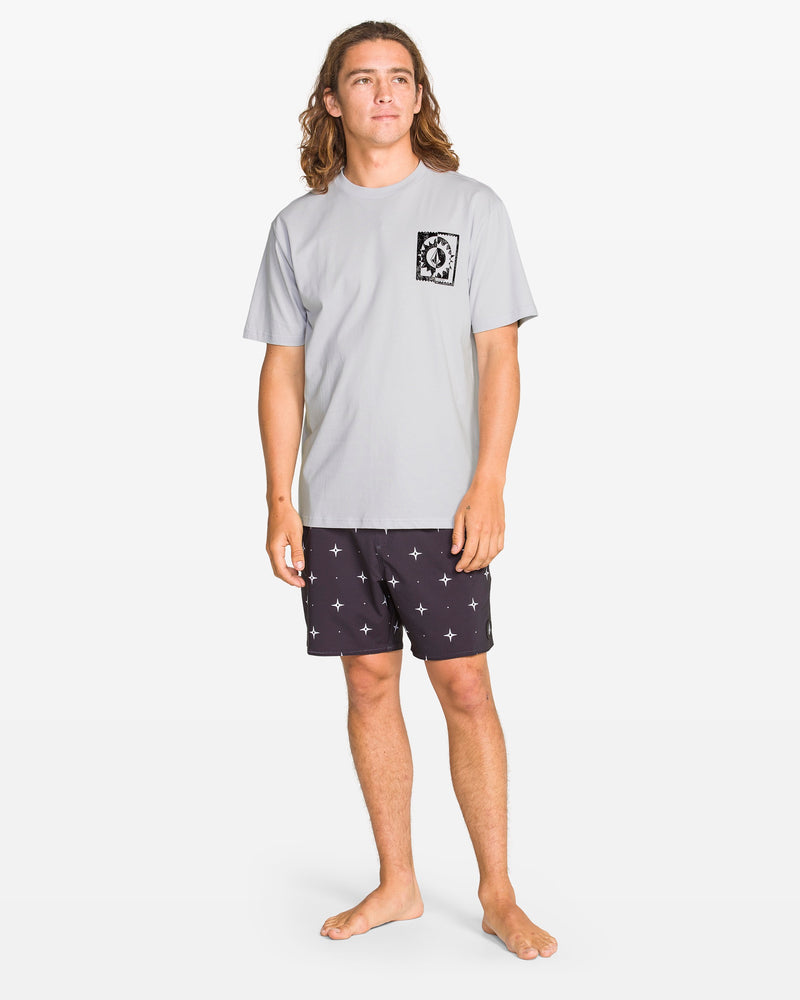 A person with long brown hair stands barefoot against a white background, wearing Volcom’s Men's Burner Short Sleeve Tee in light gray and black shorts with white star patterns.