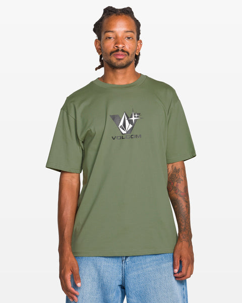 A man with dreadlocks wears the Volcom Men's Be Kind Rewind Short Sleeve Tee in olive green and light blue jeans, standing against a plain white background.