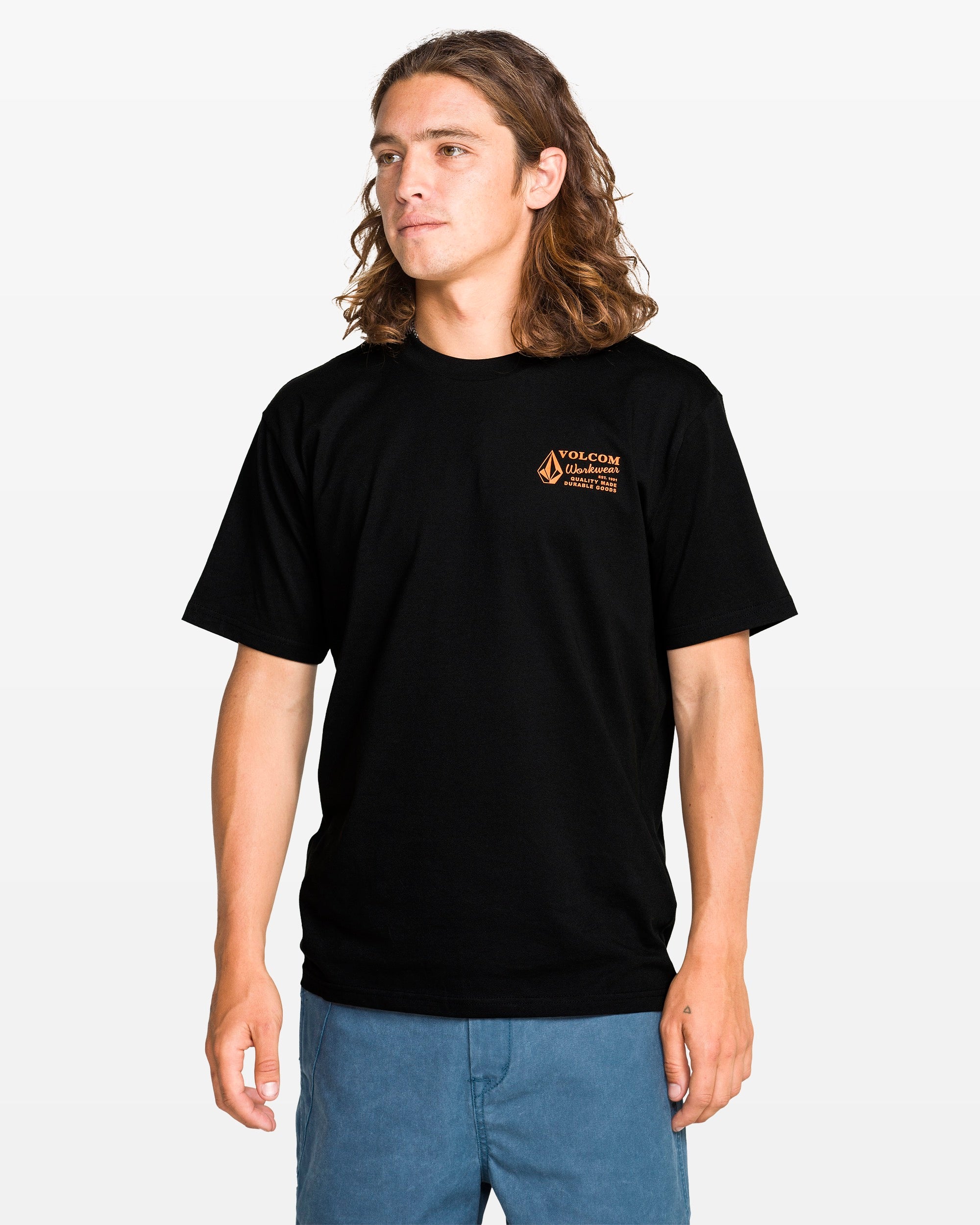 A young man with long brown hair wears a Volcom Men's Workwear Short Sleeve Tee made from organic cotton and blue shorts, standing facing forward against a white background.