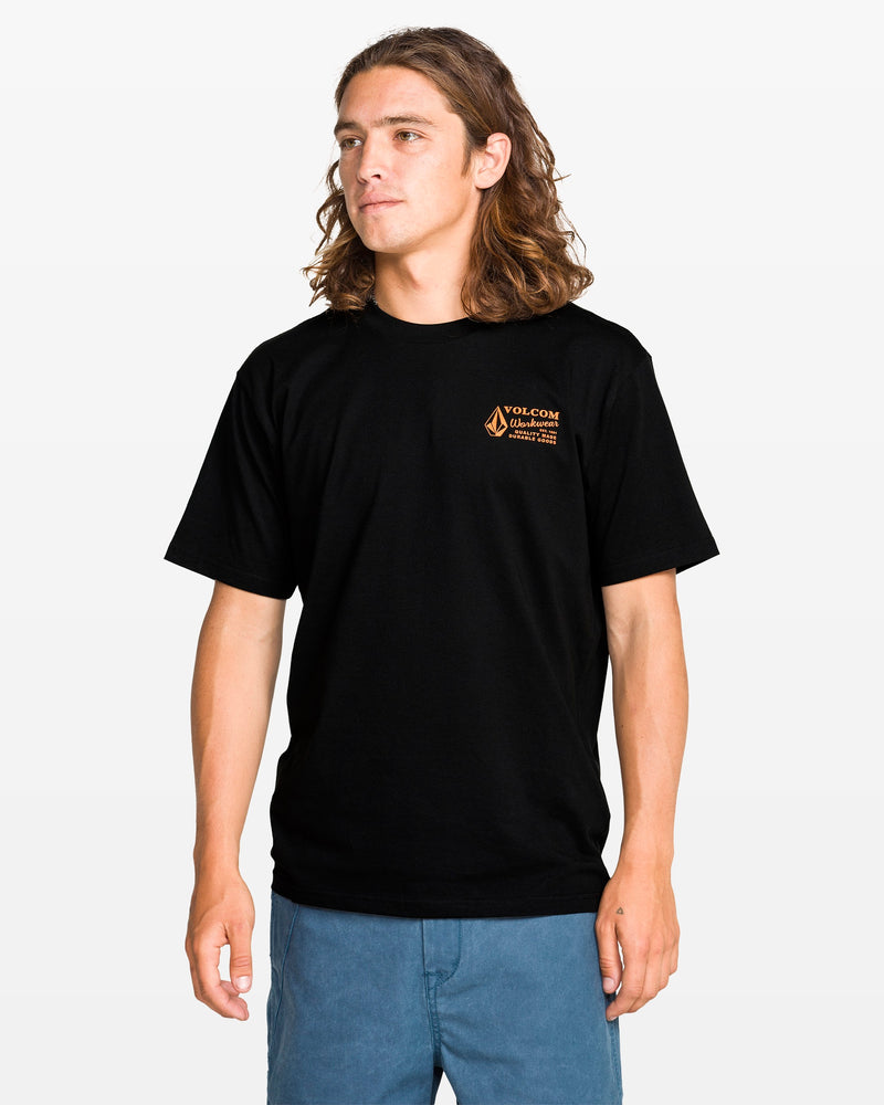 A young man with long brown hair wears a Volcom Men's Workwear Short Sleeve Tee made from organic cotton and blue shorts, standing facing forward against a white background.