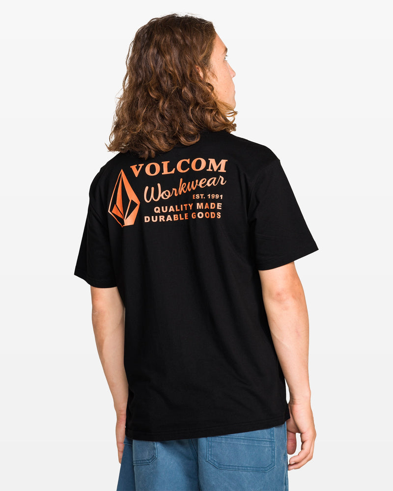 A person with long brown hair wears a Men’s Workwear Short Sleeve Tee by Volcom in black with orange lettering and back logo, paired with blue pants, against a plain light background.