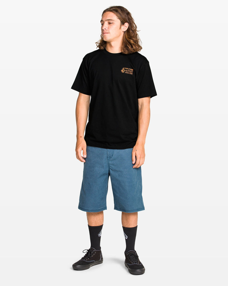 A young person with long brown hair wears the Volcom Men's Workwear Short Sleeve Tee in black, paired with blue knee-length shorts, black socks, and black sneakers, standing against a plain white background and looking to the side.