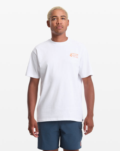 A person with short, light blonde hair wears the Volcom Men's Workwear Short Sleeve Tee in white—featuring a small orange logo and text on the upper left chest—and blue shorts, standing against a white background.