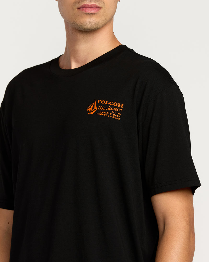 A person stands against a light background wearing the Volcom Men's Workwear Short Sleeve Tee in black, featuring a crew neck and a small orange Volcom Workwear logo with text on the left chest.