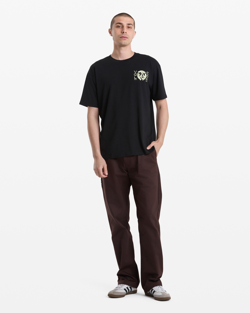 A person stands against a white background wearing the Volcom Men's Handeling Short Sleeve Shirt, brown pants, and gray Adidas sneakers. One hand is in their pocket and they have a neutral expression.