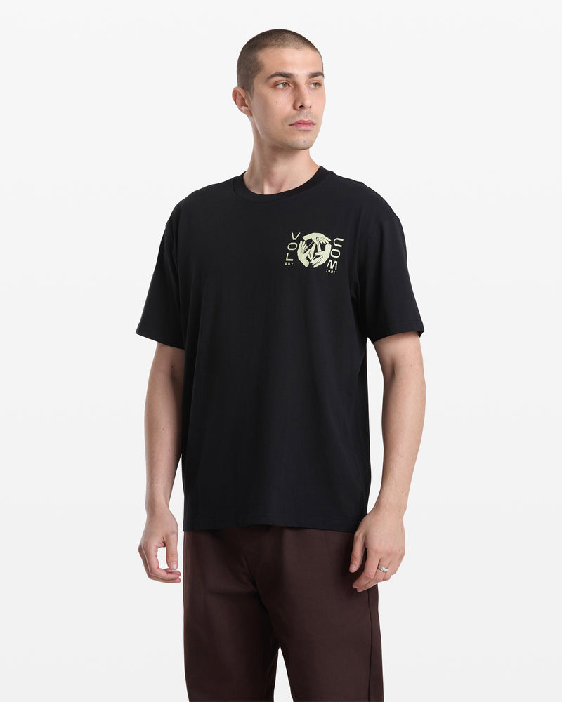 A man with short hair wears the Volcom Men's Handeling Short Sleeve Shirt, a black 100% cotton tee with a yellow geometric graphic and text on the left chest, paired with brown pants, against a plain white background.