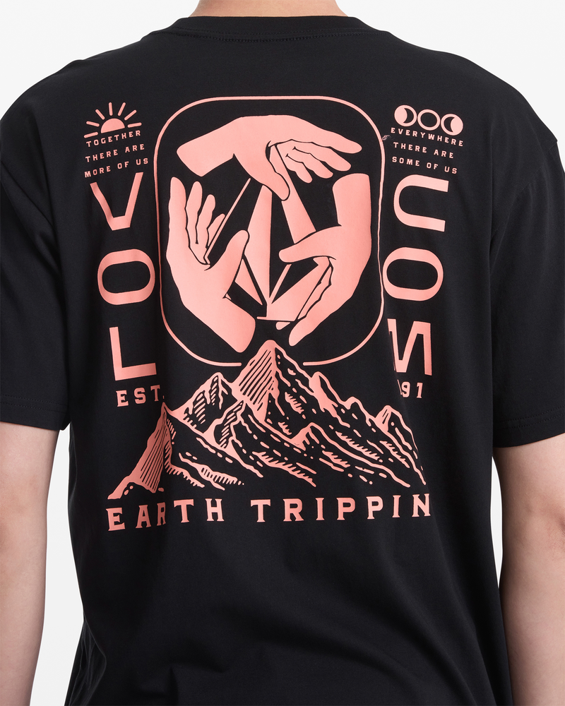 A person wears the Volcom Men's Handeling Short Sleeve Shirt, a 100% cotton slim tee with pink hand graphics above a mountain, plus "VOLCOM," "Earth Trippin," "Outdoor Earth Tripper EST. 1991," and motivational phrases.