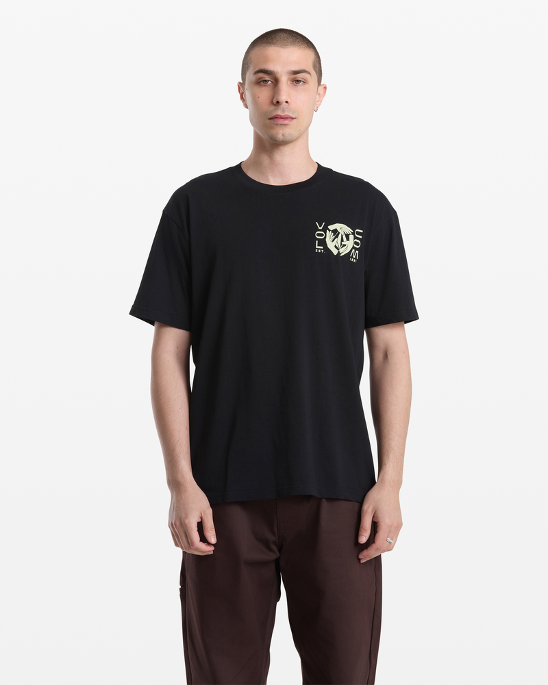 A man with short hair wears the Volcom Men's Handeling Short Sleeve Shirt in black 100% cotton with a small yellow graphic and text on the left chest, paired with brown pants against a plain white background.