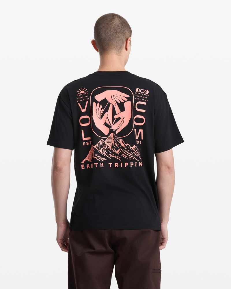 A person with short hair, seen from behind, wears the Volcom Men's Handeling Short Sleeve Shirt made of 100% cotton, featuring a large pink graphic of abstract hands above mountain peaks with "VOLCOM" and "Outdoor Earth Tripper" on the back.