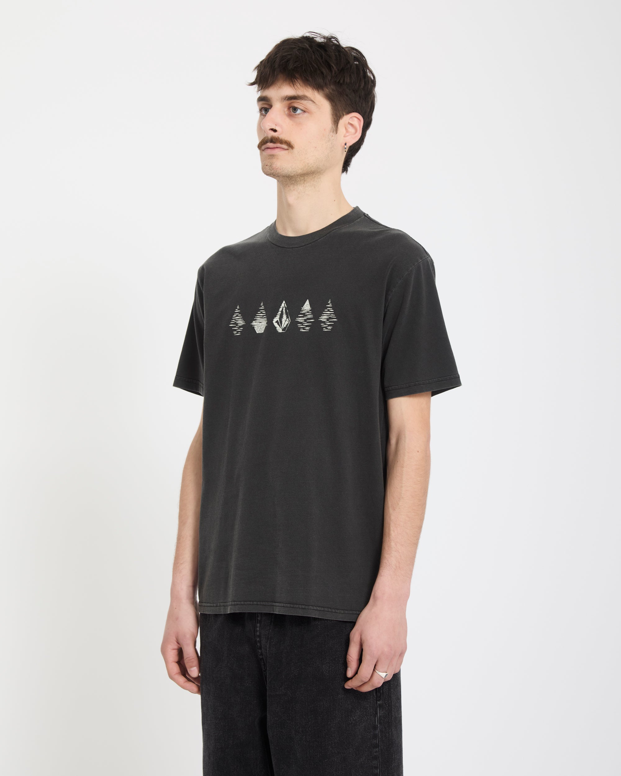 A young man with short dark hair and a mustache stands against a white background, wearing Volcom’s Men's Lunar Phases Short Sleeve Tee in black, featuring a row of white abstract designs on the chest, and black jeans. He looks slightly to the side.