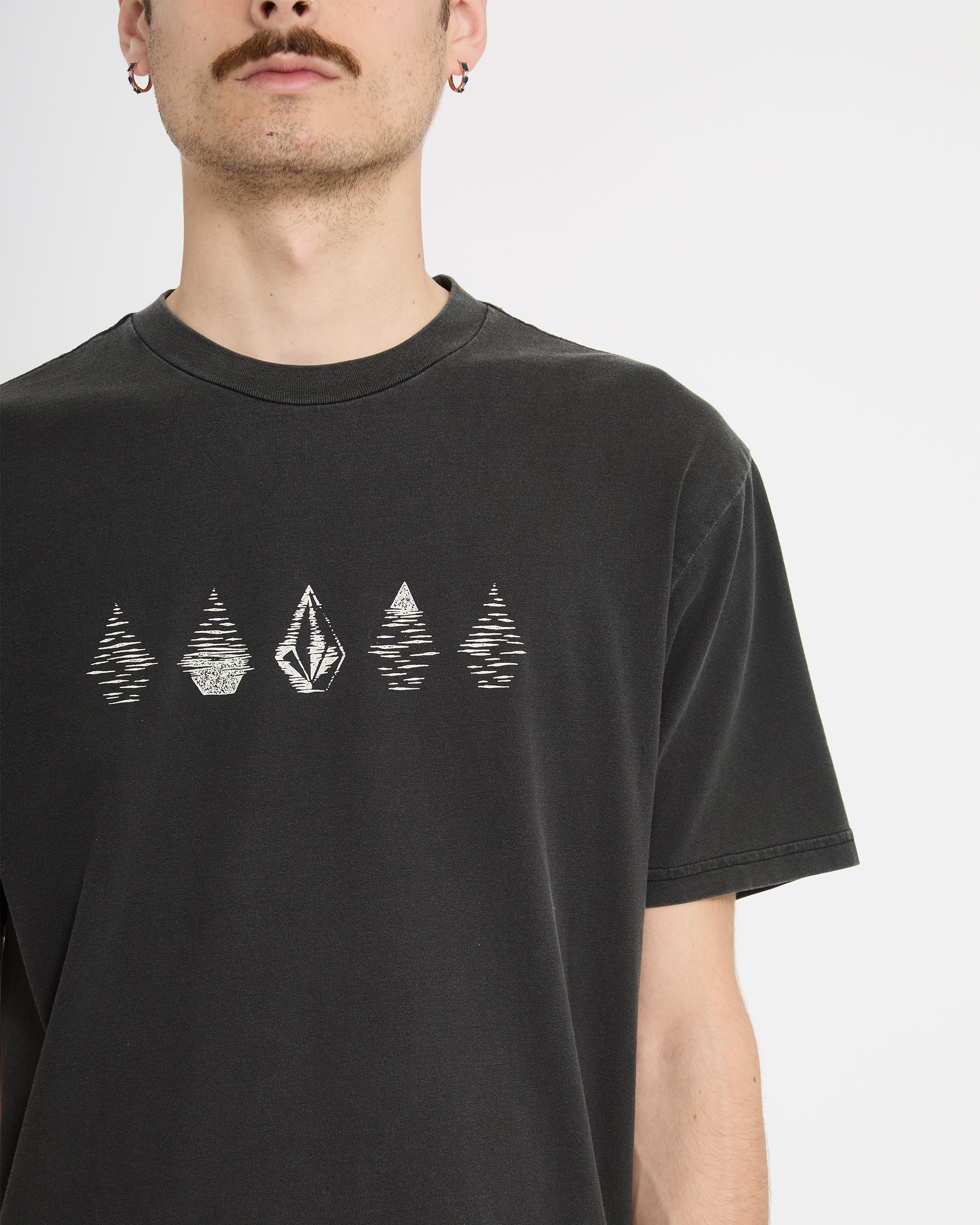 A man with a mustache and earrings wears the Volcom Men's Lunar Phases Short Sleeve Tee, featuring geometric tree and diamond graphics on black, photographed against a plain white background.