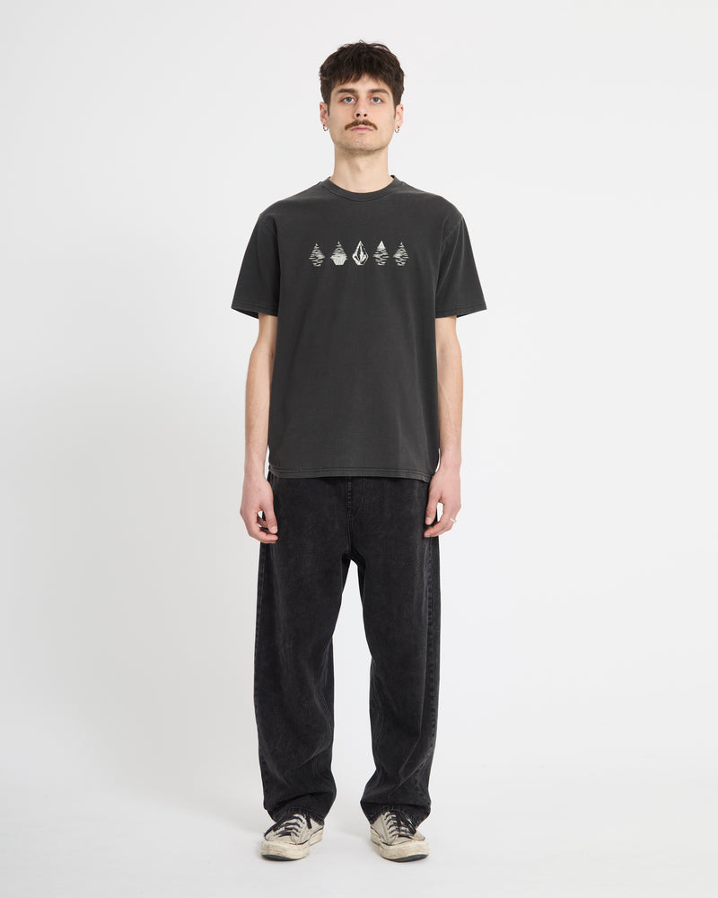 A person stands facing forward against a plain white background, wearing Volcom's Men's Lunar Phases Short Sleeve Tee, loose black jeans, and white sneakers.