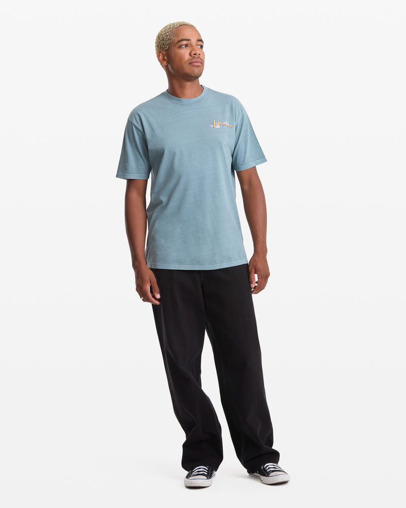 A person with short blond hair wears a Volcom Men's Zorch Short Sleeve Tee in light blue, paired with black pants and black-and-white sneakers, standing against a plain white background.