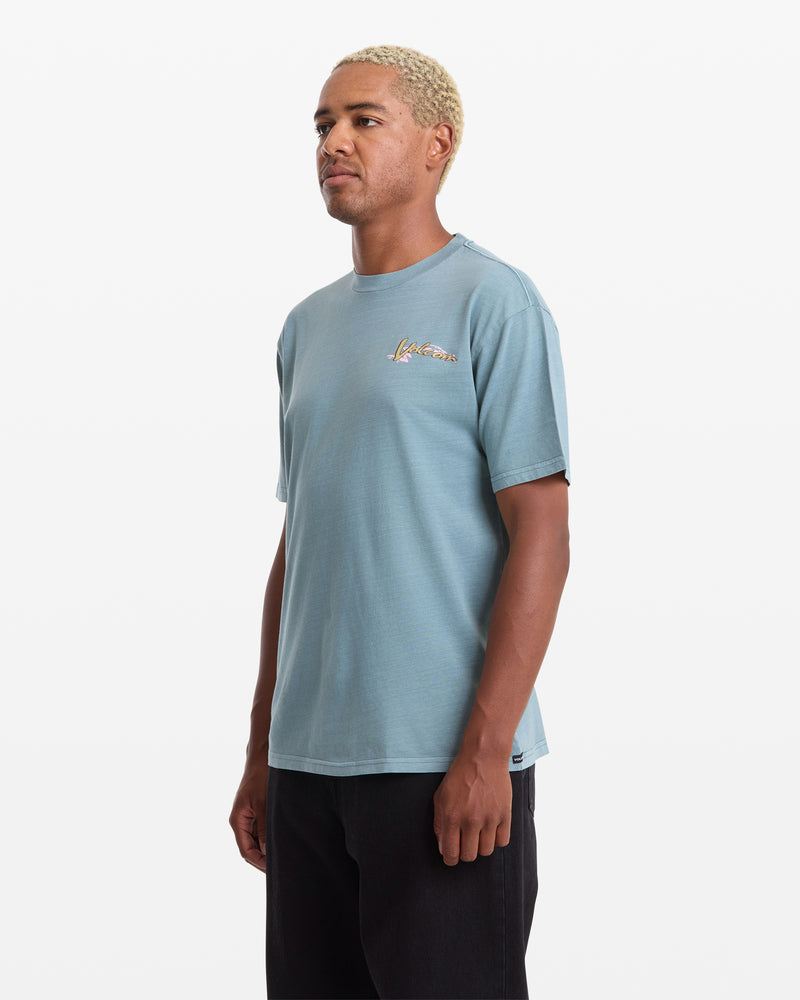 A man with short blond hair wears the Volcom Men's Zorch Short Sleeve Tee in light blue 190gm cotton, featuring a small chest graphic, paired with black pants and standing against a plain white background.