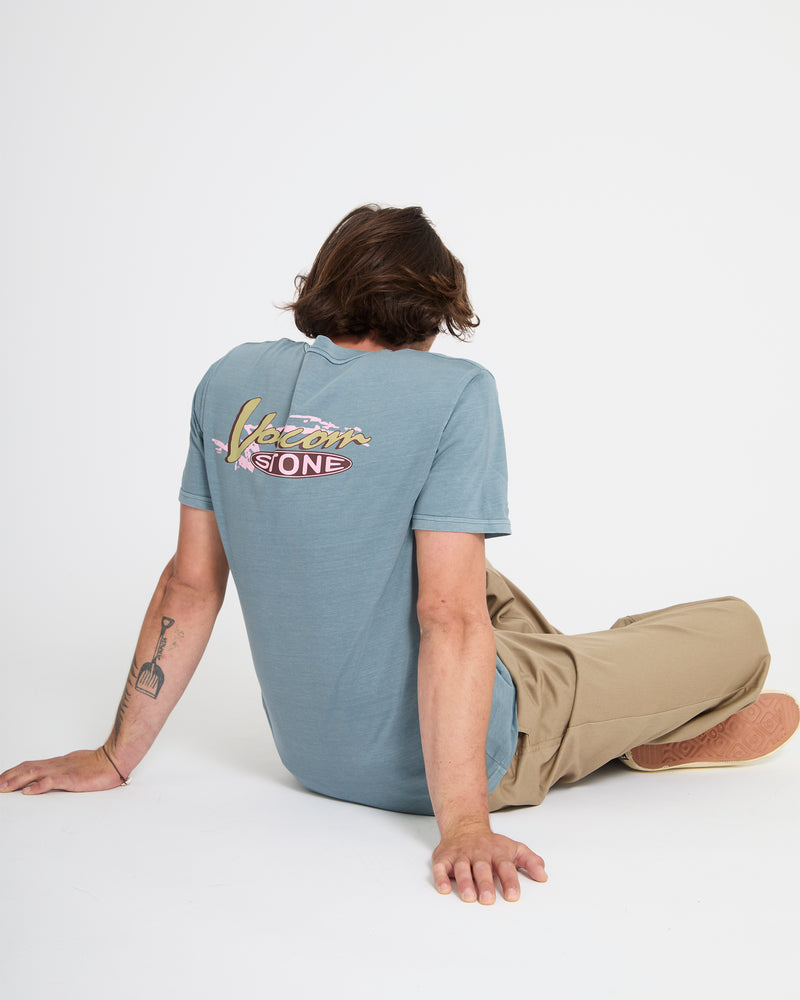 A person with shoulder-length brown hair sits on the floor facing away, wearing a loose-fit light blue Volcom Men's Zorch Short Sleeve Tee and beige pants. Their left arm, showing a guitar tattoo, is extended behind them for support.