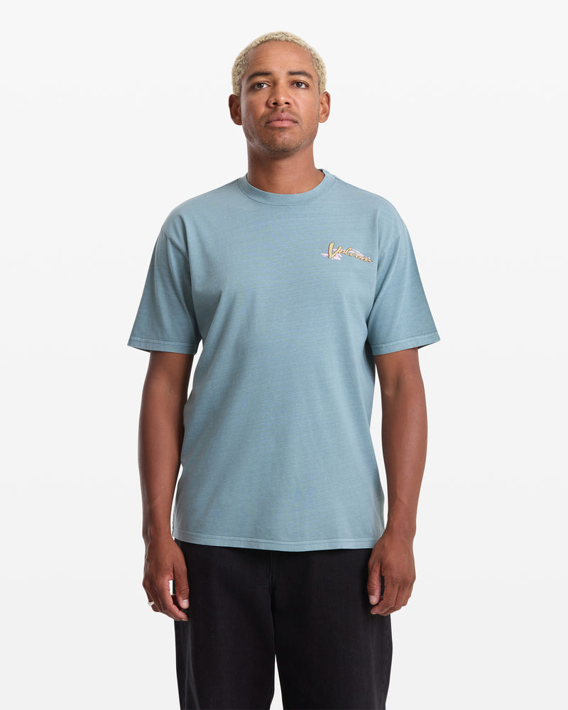 A person with short, bleached hair wears the Volcom Men's Zorch Short Sleeve Tee in light blue, a loose fit 190gm cotton shirt with a small colorful chest graphic, standing against a plain white background.