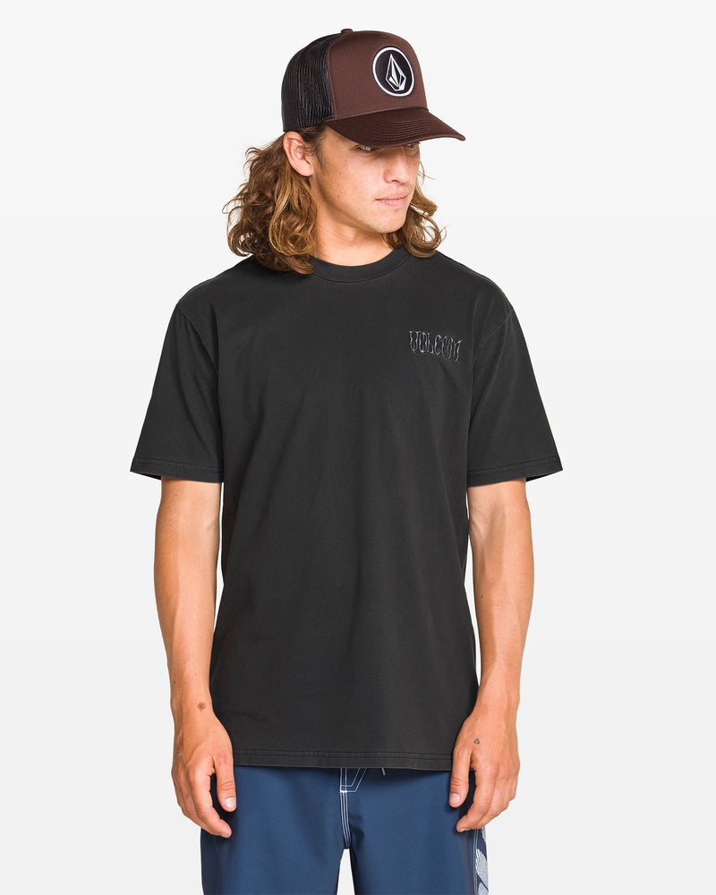 A man wearing the Volcom Men's Proto Type Short Sleeve Tee, blue shorts, and a black Volcom cap stands against a white background, looking down and to the side, channeling effortless style.