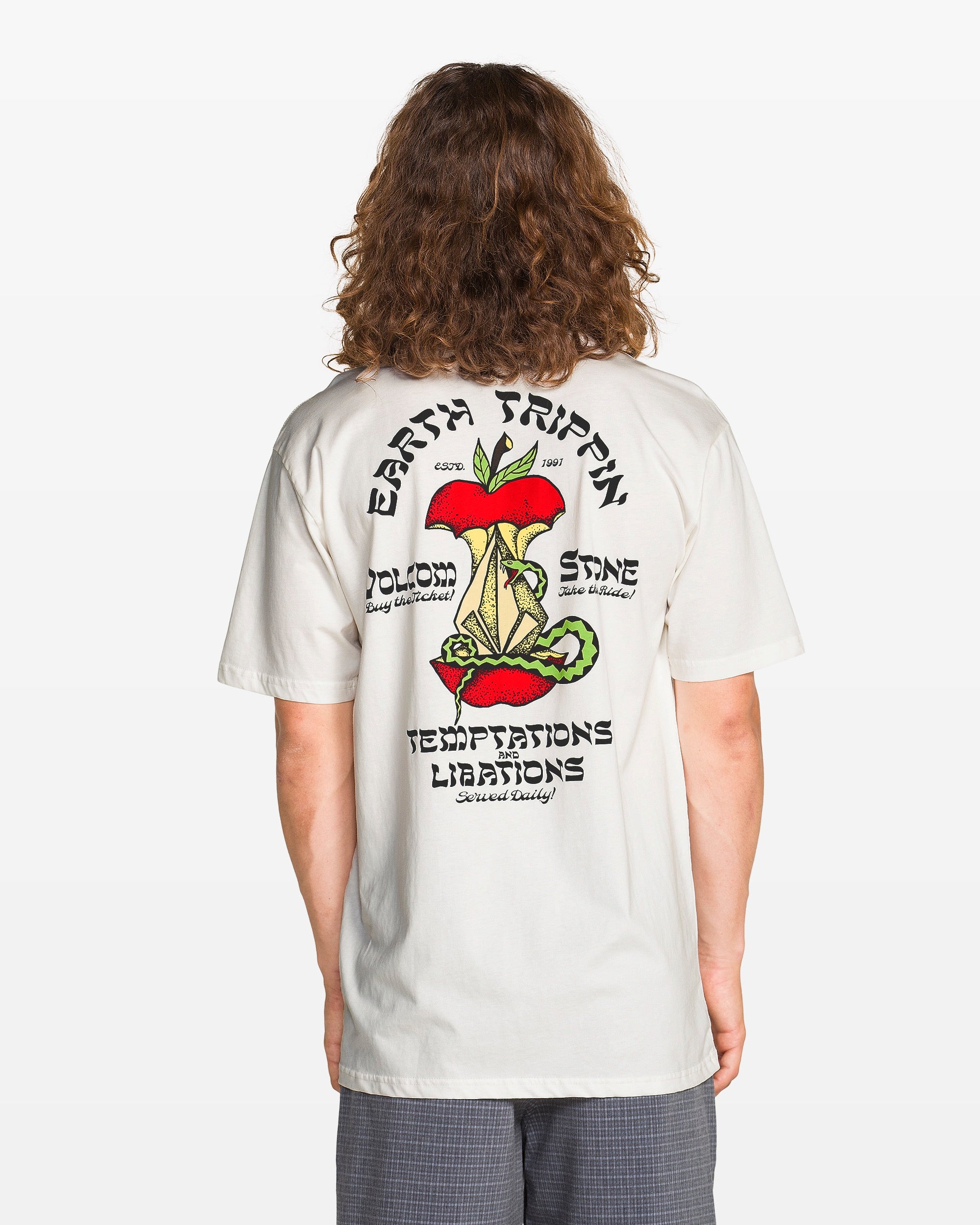 A person with long hair wears the Volcom Men's Temptations Short Sleeve Tee, a soft white cotton crewneck featuring a colorful mushroom and plant graphic on the back, plus the words: EARTH TRIPPIN, BLOOM, SHINE, TEMPTATIONS, LIBATIONS.
