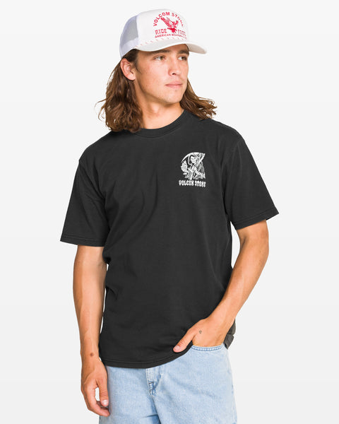 A man with long brown hair, wearing a white trucker hat, light blue jeans, and the Volcom Men's Eternal Short Sleeve Tee in black with a soft-hand screenprint, stands against a plain background with one hand in his pocket and looks to the side.