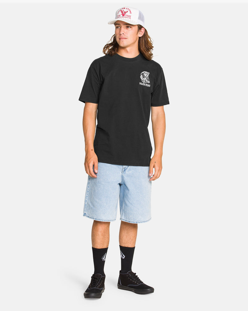 A young man with long hair wears a white cap, Volcom Men's Eternal Short Sleeve Tee in black, light blue knee-length denim shorts, black socks, and black sneakers, standing against a plain white background.