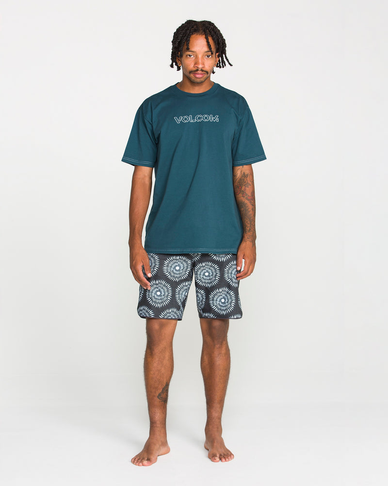 A man stands barefoot against a white background, wearing Volcom's Men's Crassy Short Sleeve Tee in teal—crafted from premium cotton with a soft-hand screenprint—and patterned black-and-white shorts.