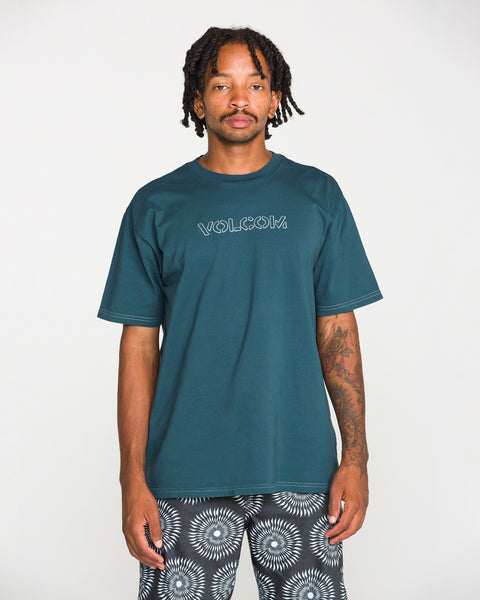 A man with short dreadlocks stands against a plain white background, looking directly at the camera, wearing the Volcom Men's Crassy Short Sleeve Tee in premium cotton with a teal graphic, paired with patterned shorts.