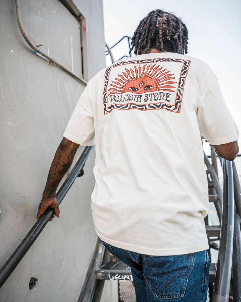 A person with braided hair, walking up metal stairs, is wearing the Volcom Men’s Helios Short Sleeve Tee—a 190g cotton premium shirt featuring a large sun graphic and VOLCOM STORE on the back in soft-hand screenprint.