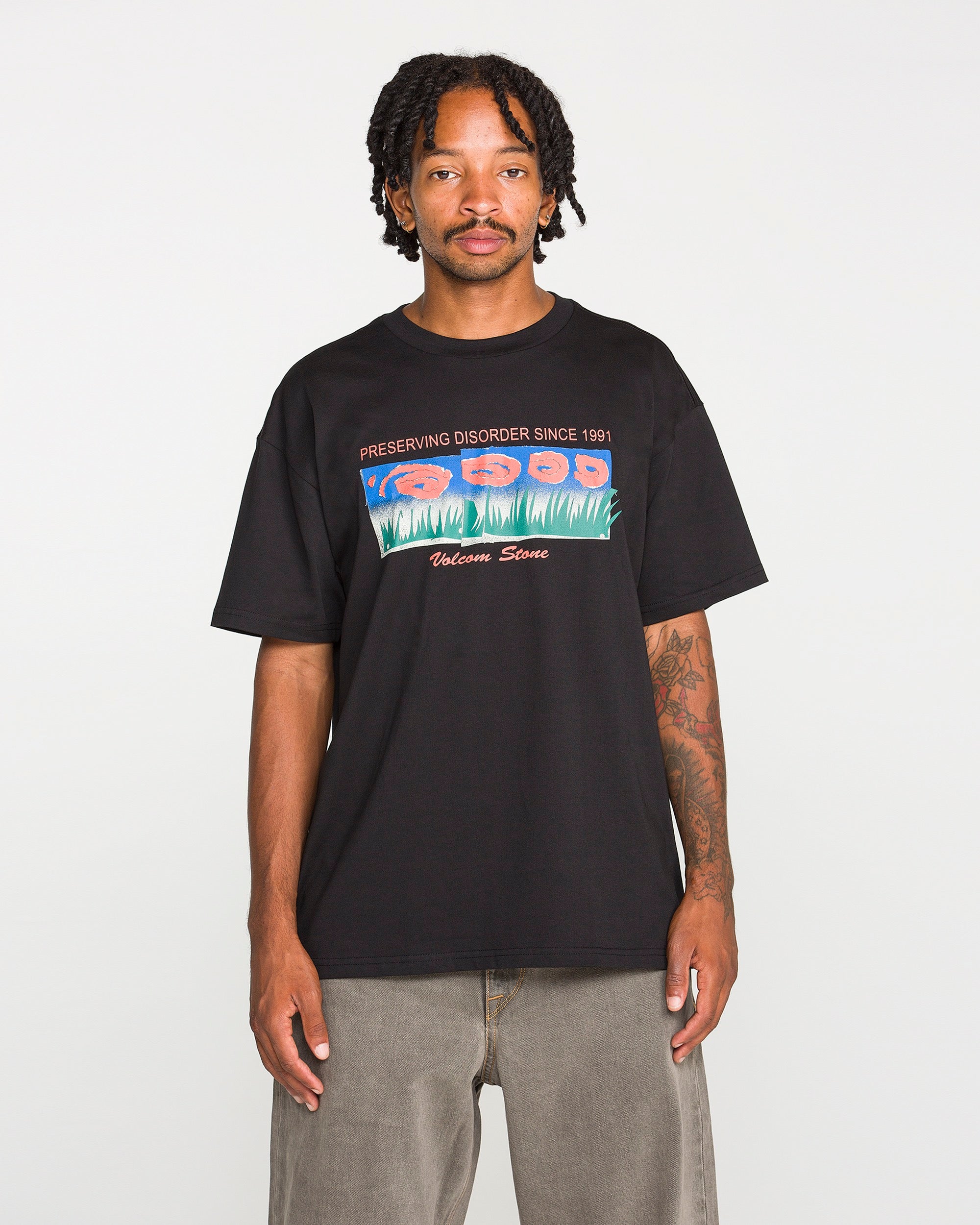 A man stands facing forward against a plain white background, wearing the Volcom Men's Featured Artist Antonio Carrau Short Sleeve Tee—a premium black shirt adorned with colorful graphics and the text "PRESERVING DISORDER SINCE 1991" and "Welcome Stone.