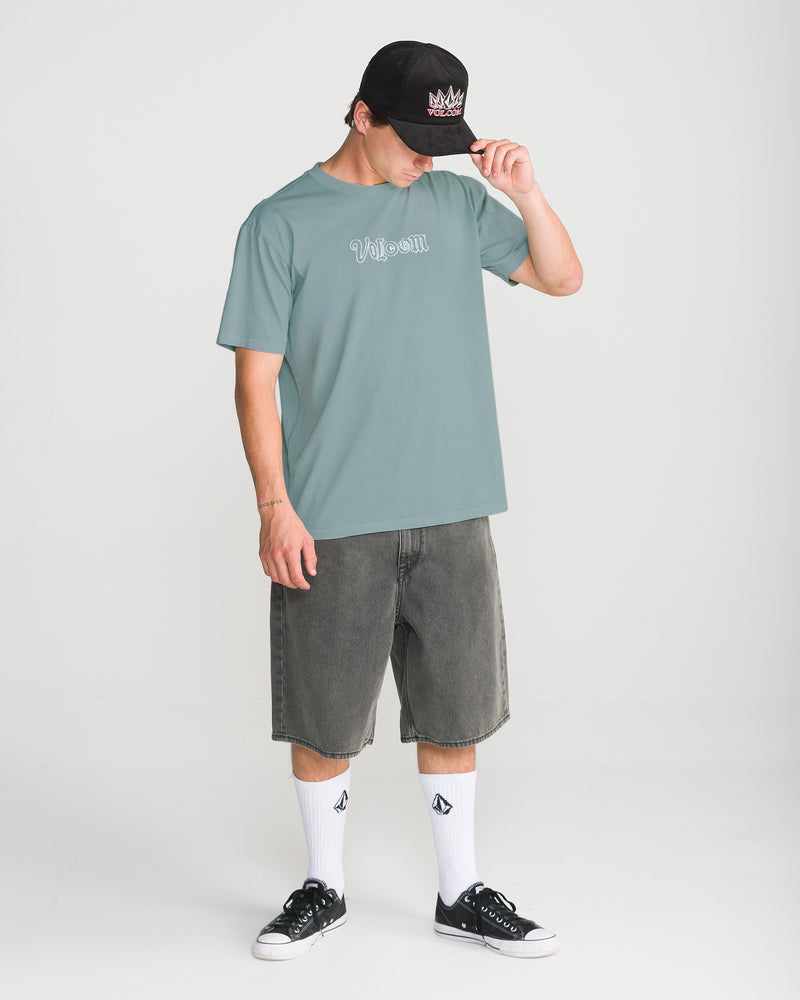 A person wearing a dusty blue Volcom Men’s Ransom Stitch Short Sleeve Tee with an embroidered logo on the chest, grey denim shorts, white crew socks, and black sneakers.