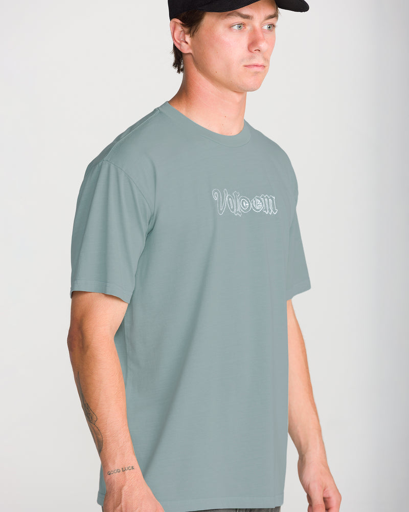 A man wearing a sage green, loose fit Men’s Ransom Stitch Short Sleeve Tee by Volcom with a white outlined embroidered logo on the chest and a black baseball cap.
