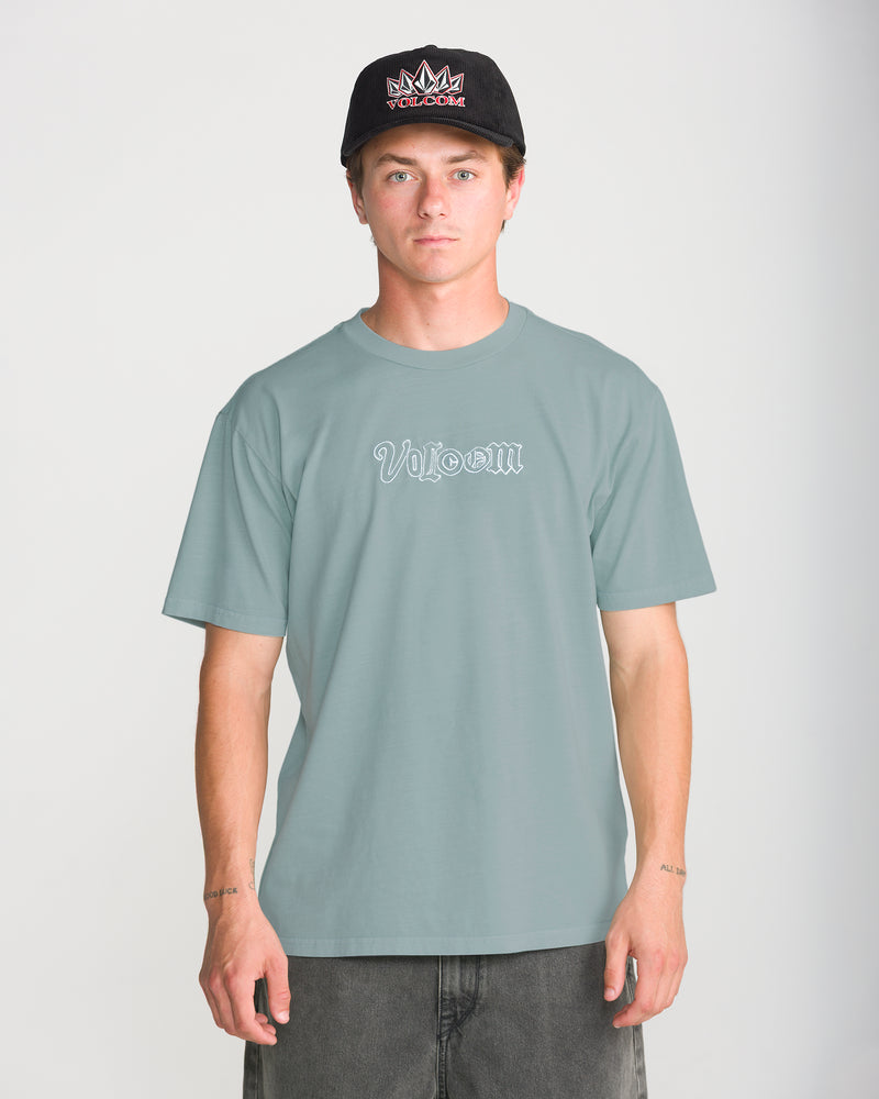A man wears a grey, 100% cotton Volcom Men’s Ransom Stitch Short Sleeve Tee with an embroidered white script logo on the chest and a black baseball cap.
