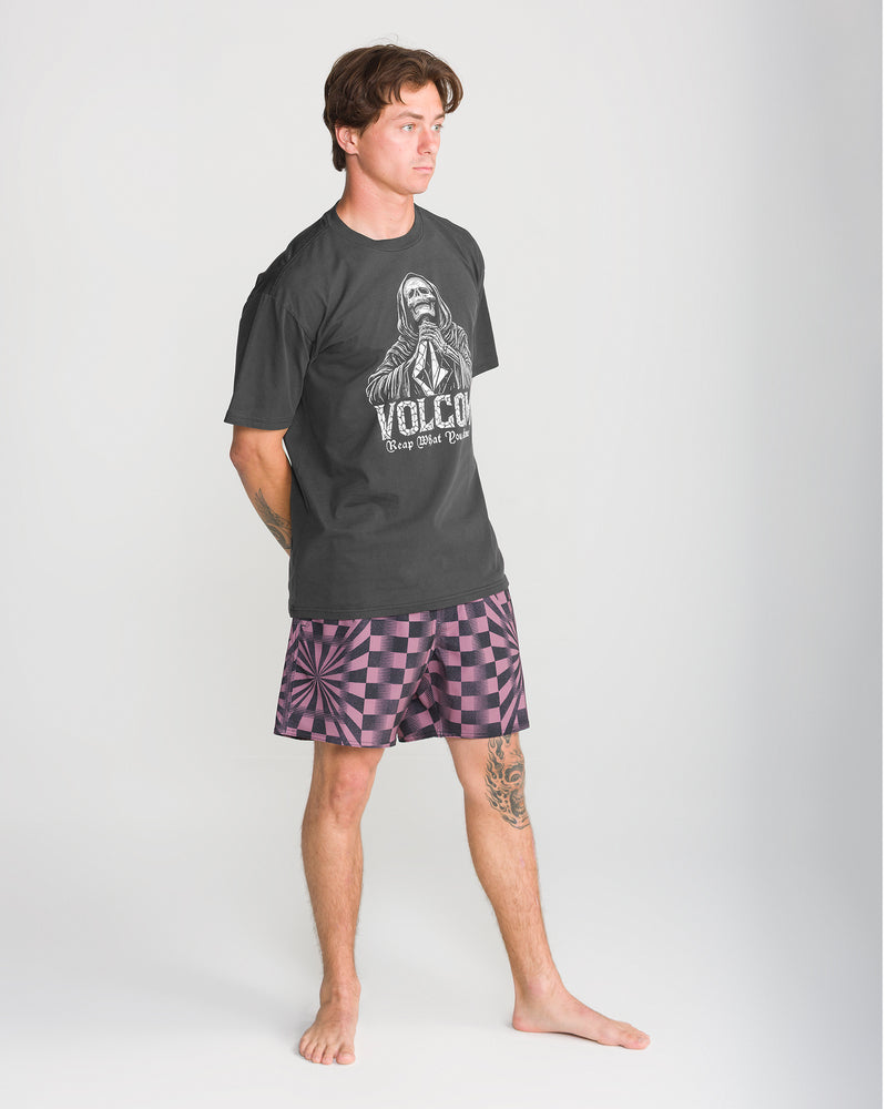 A man wears a dark gray Volcom Men’s Reap What You Stone Short Sleeve Tee with a skeleton graphic and purple checkered shorts.