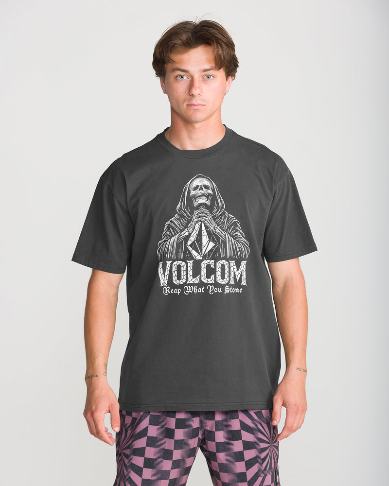 A man wearing a charcoal grey Men’s Reap What You Stone Short Sleeve Tee featuring a skeleton in a hooded robe with hands pressed together above the text Reap What You Stone.