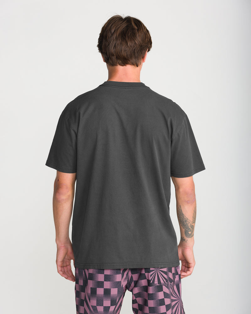 Back view of a person wearing a plain dark grey Men’s Reap What You Stone Short Sleeve Tee by Volcom and patterned shorts.