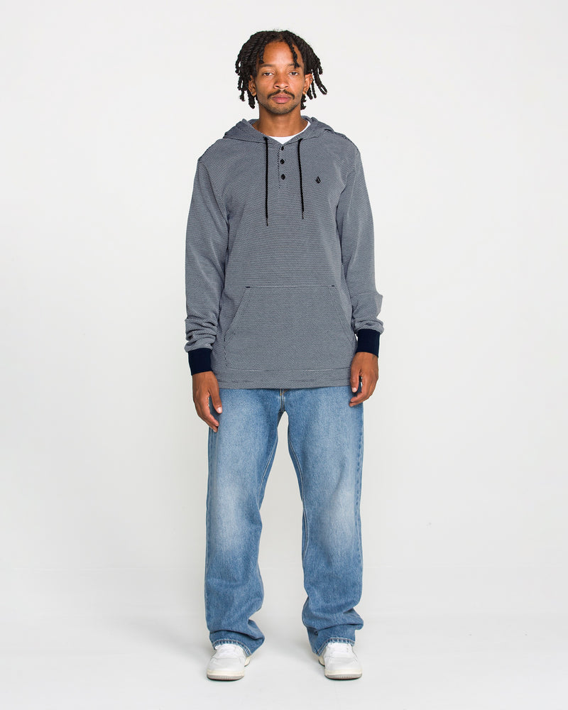A man with short dreadlocks stands against a plain white background, wearing the Volcom Men's Rowan Thermal Long Sleeve Shirt for midweight warmth, paired with light blue jeans and white sneakers. He looks directly at the camera with a neutral expression.