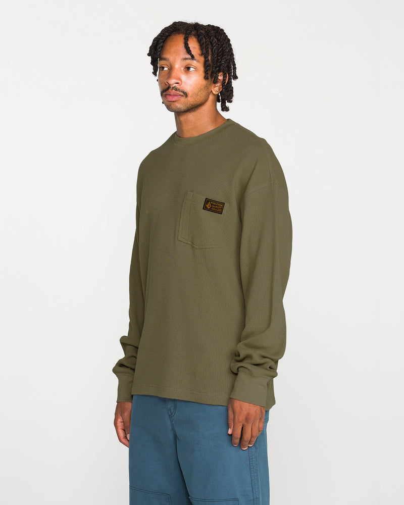 A man wearing a Volcom Men's Overtime Workwear Thermal Long Sleeve Shirt in olive green, featuring a chest pocket, and blue pants stands against a plain white background. Made from a cotton knit fabric, this thermal shirt provides comfort and durability for everyday wear.