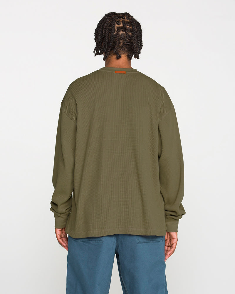 A person with braided hair, wearing the Volcom Men's Overtime Workwear Thermal Long Sleeve Shirt in olive green and blue pants, stands facing away from the camera against a plain white background.