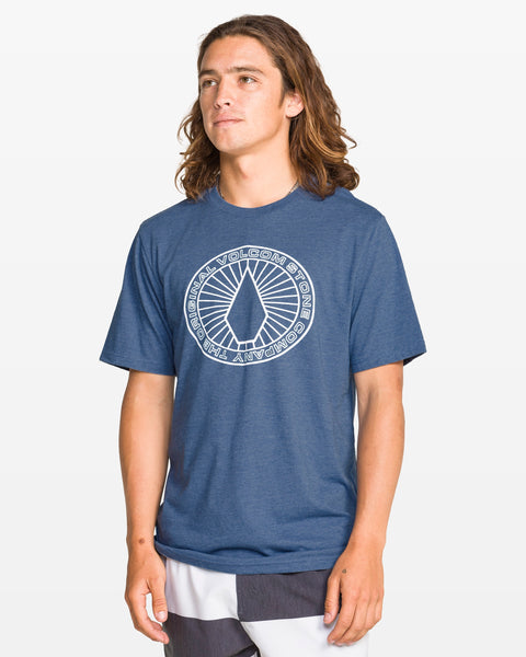 A young man with long brown hair wears a Volcom Men's Men blue crew neck short sleeve tee featuring a white circular logo on the chest and black-and-white shorts, standing against a plain white background.