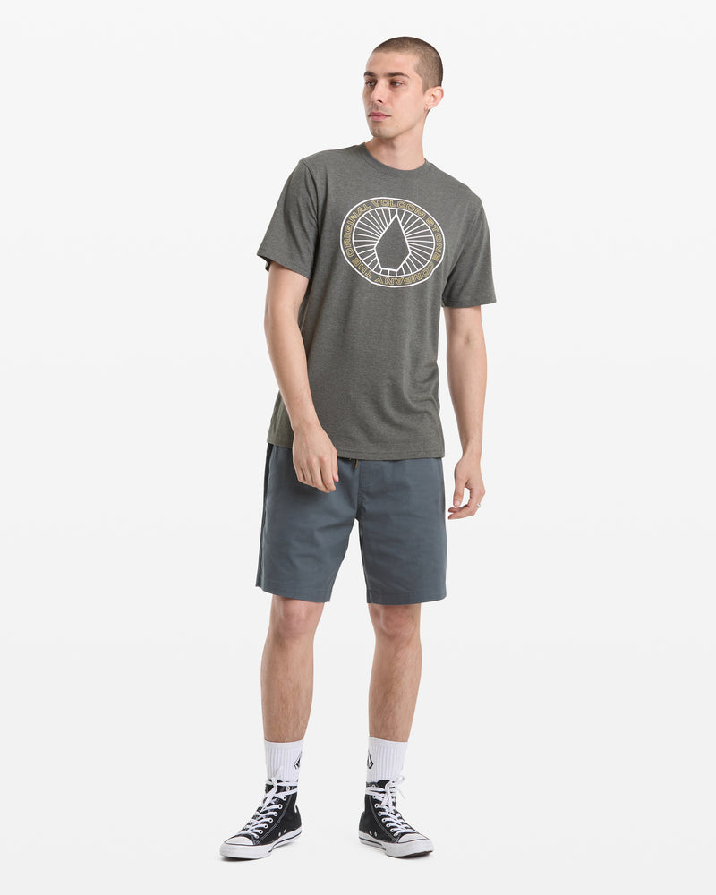A man stands against a white background wearing the Volcom Men's Medalist Short Sleeve Tee, blue-gray shorts, white crew socks, and black high-top sneakers. He looks off to the side with a relaxed posture.