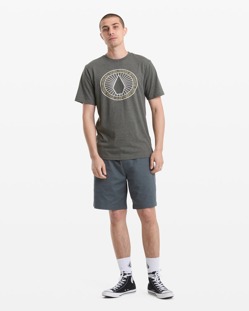 A young man with short hair stands against a white background wearing a Volcom Men's Medalist Short Sleeve Tee, blue-gray shorts, white socks, and black high-top sneakers. One hand is in his pocket.
