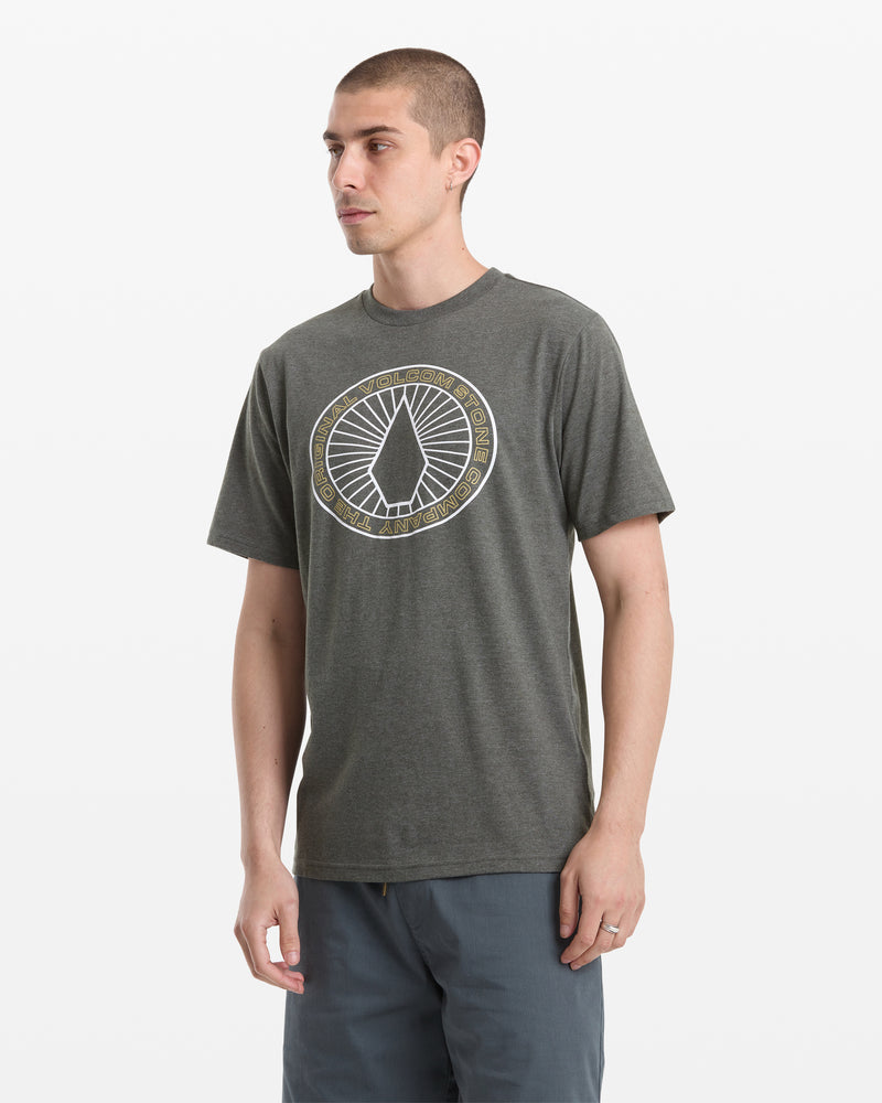 A man wearing the Volcom Men's Medalist Short Sleeve Tee, a dark gray heather shirt with a circular chest graphic, and blue-gray shorts stands against a plain white background.