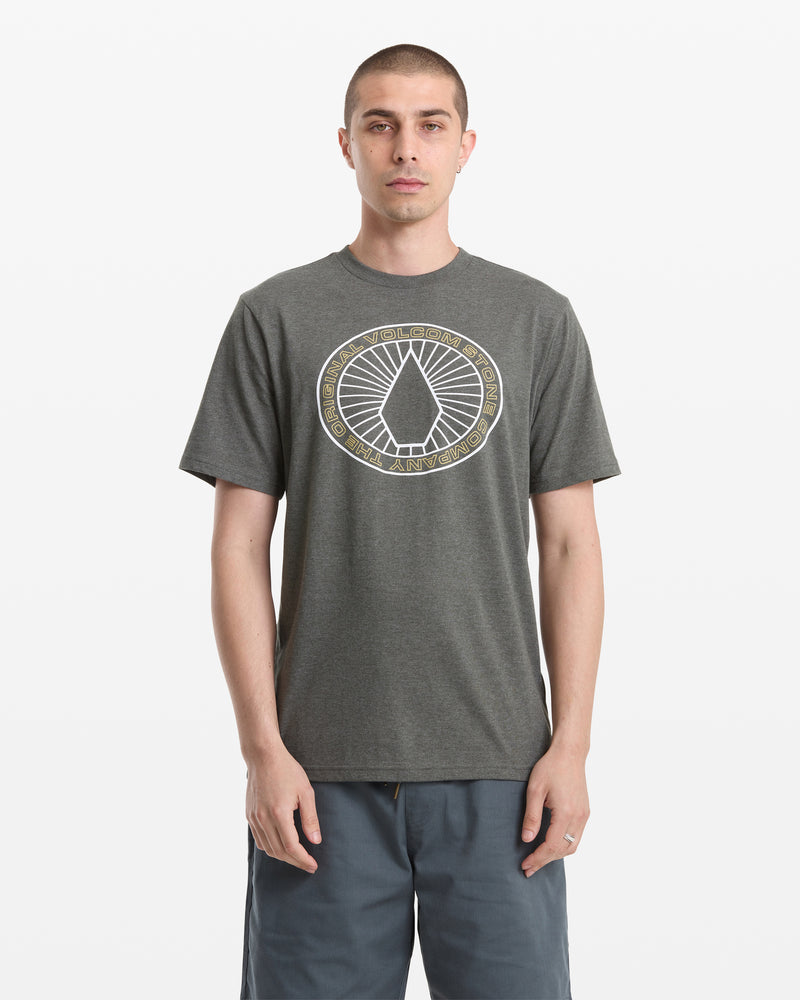 A man faces forward wearing a Volcom Men's Medalist Short Sleeve Tee in dark gray heather, featuring a circular geometric logo and text on the chest, paired with gray shorts. The backdrop is plain white.
