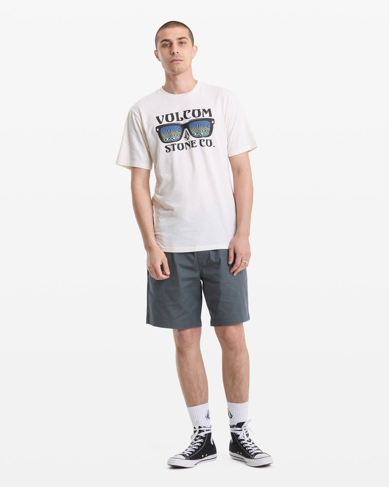 A man stands against a white background wearing a Volcom Men's Sunnies Short Sleeve Tee with a sunglasses graphic, gray shorts, white crew socks, and black high-top Converse sneakers.